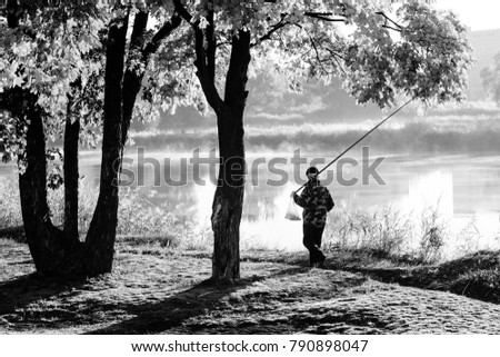 Autumn landscape in city park. Fisherman walking on path along foggy river. Trees on river bank. Black and white photo
