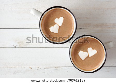 Two cups of hot chocolate with heart shaped marshmallows over a white wood background