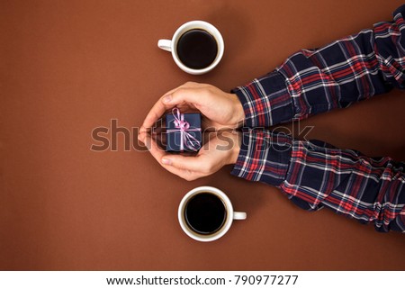The male hands are closed on a brown background. The guy gives a gift to a girl. Men's hands next to two cups of coffee and with gift.