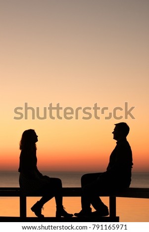 Vintage look image of young couple sitting and watching at each other near the ocean during the sunset. Silhouette picture of couple at the sunset.