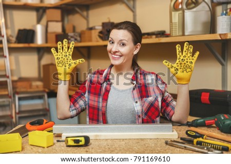 Beautiful smiling caucasian young brown-hair woman in plaid shirt, gray T-shirt, yellow gloves working in carpentry workshop at wooden table place with piece of iron and wood, different tools