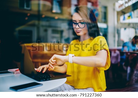 Young businesswoman in eyeglasses looking at display of wristwatch and managing time while checking email on laptop.Freelancer with smartwatch on hand working on article at netbook before deadline Royalty-Free Stock Photo #791227360