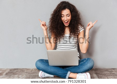 Nice woman with beautiful smile email with her friend using silver laptop while sitting with legs crossed on the floor, being isolated over grey background