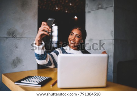 Cheerful beautiful afro american female making selfie on front camera of telephone for publishing in social networks sitting at laptop computer in campus.Hipster girl taking photos on smartphone