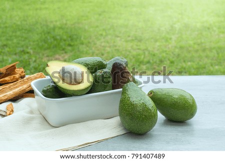 Avocado and cut avocado in white bowl on light blue wood against green field