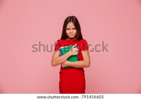 Unhappy displeased girl in red dress holding book and looking camera isolated over pink