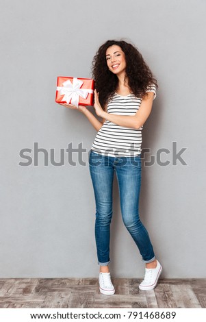 Full size picture of cheerful pretty woman holding gift-wrapped present with white bow being isolated over grey wall