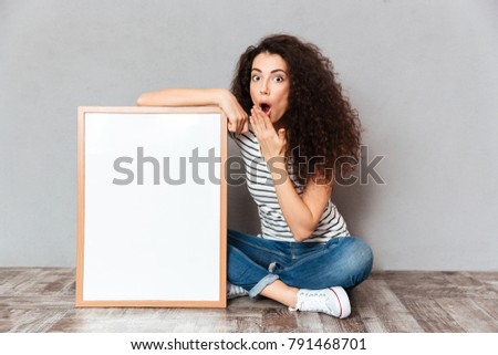 Funny portrait of adult girl in jeans sitting in lotus pose on the floor expressing embarrassment and surprise, while holding picture in frame copy space