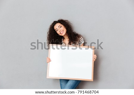 Caucasian woman with beautiful hair posing over grey wall holding piece of art in hands, expressing admiration about portrait copy space
