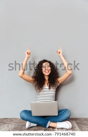 Picture of agitated woman sitting with legs crossed on the floor being happy and excited, putting index fingers in the air over grey wall