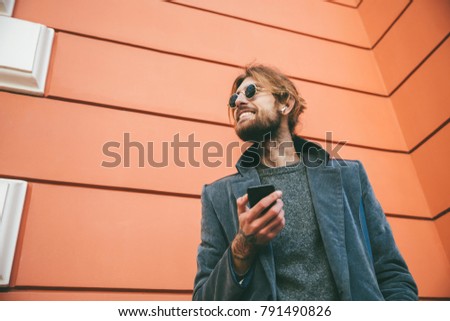Portrait of a happy bearded man dressed in coat standing against a wall on a city street and holding mobile phone