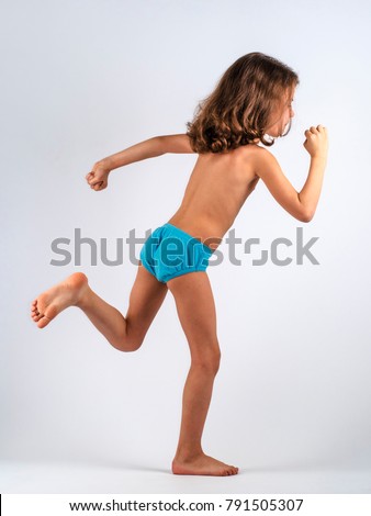 Cute boy with long curly hair in swimming trunks, performing gymnastic exercises. Gray background.