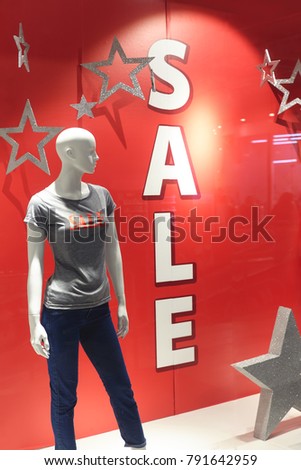 A mannequin in a gray T-shirt and jeans is on display in a shop window with an inscription of a sale.