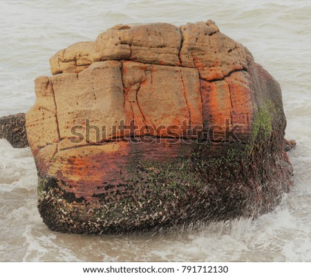 Colourful large brown and orange rock with wave crashing around it in the sea