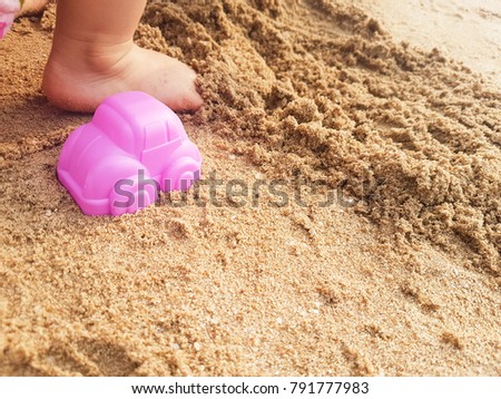 Pink car model for playing at the beach with the sand. There are feet of children in the picture.