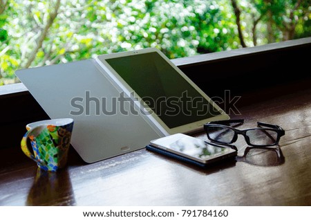 Working to connection with tablet computer , smartphone and a coffee cup with glasses on wood table with green tree natural in front of office.Concept relax from work.