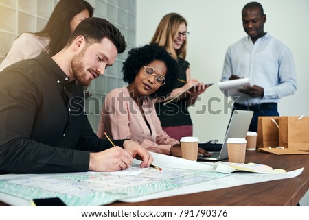 Two designers discussing building blueprints together while working at a table in an office with colleagues in the background