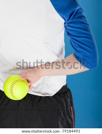 A woman in a blue and white baseball jersey holds a softball behind her back, preparing to throw it. Photographed with studio lights in front of a blue backdrop.