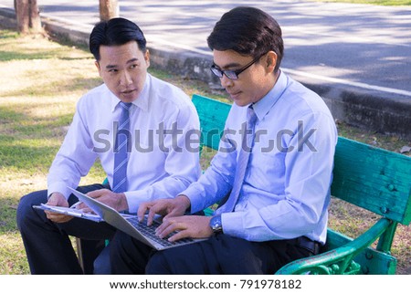 Businessman, They are sitting on bench in park. He is play notebook and  search internet. Another one holding paper and talking about business in  relax time. Photo concept  business and relax time.