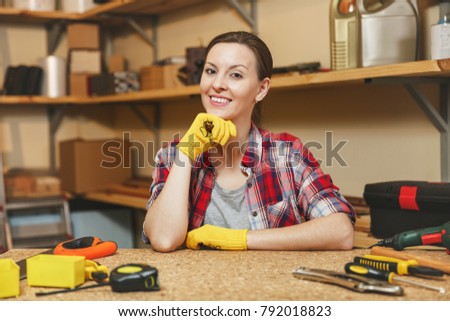 Beautiful smiling caucasian young brown-hair woman in plaid shirt, gray T-shirt, yellow gloves working in carpentry workshop at wooden table place with different men's work tools. Gender equality