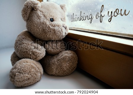 The sad teddy bear sits on the windowsill and looks sadly out the window, it's raining, the glass is covered with drops. Handwritten inscription on a glass " thinking of you"