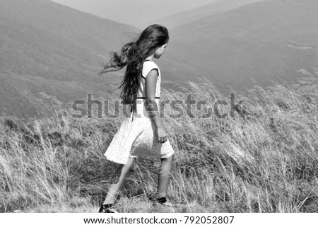 Back viev of young little brunette girl in white lace summer dress walking in mountain valley with deep dry spikelet grass in spring sunny day outdoor on natural blue background, horizontal picture