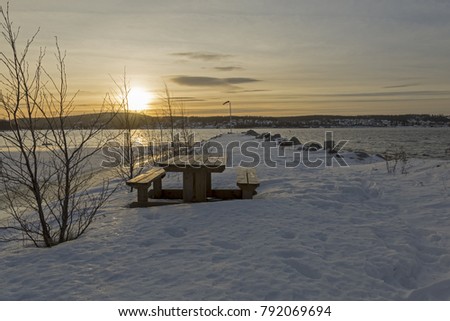 Pier in winter with snow and a bench in foregground and the sunset in background, picture from Northern Sweden.