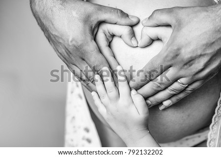 Hands of female male and baby make heart shape cute sign on bare belly of pregnant woman mother on grey background