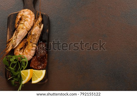 Fried tiger shrimps with lemon, sun-dried tomatoes and greens or rukkola on a dark wooden cutting board.