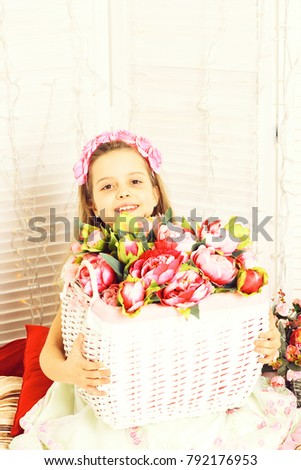 young smiling girl with wreath on curly hair and cute dress holding vintage basket of pink decorative peonies on white studio background, copy space