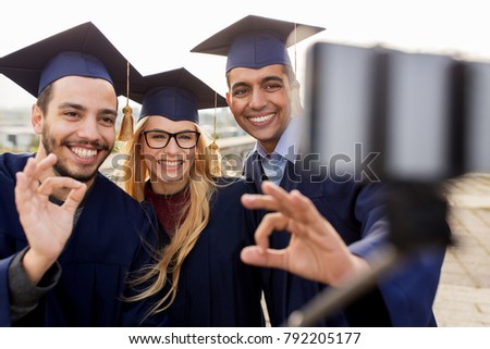 education, graduation, technology and people concept - group of happy international students in mortar boards and bachelor gowns taking selfie by smartphone outdoors and showing ok hand sign