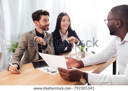 Portrait of young smiling multinational businessmen and businesswoman sitting at the table and discussing new project while working together in office 