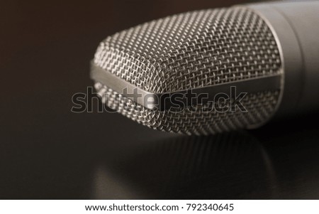 A silver condenser microphone is placed on a shiny surface. The object reflects off the black table surface.