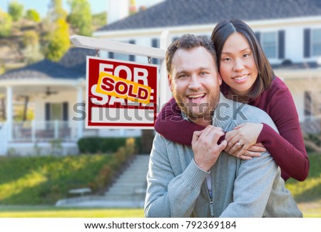Mixed Race Caucasian and Chinese Couple In Front of Sold For Sale Real Estate Sign and House.