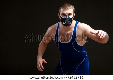 A young dark-haired man fighting Greco-roman wrestling and grappling in a blue   tights  , training mask stands before the start of the  wrestling against a black isolated  background