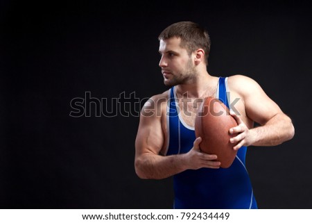 A young dark-haired male wrestler in a blue wrestling tights smiles and holds a rugby ball in the hands on a  black isolated background