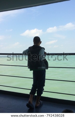 ASIAN BOY HOLDING HANDRAIL SIDE ON BOAT DURING SEA JOURNEY , BLUE SKY CLOUD OVER WATER IN BACKGROUND