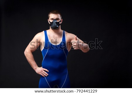 A young dark-haired man fighting Greco-roman wrestling and grappling in a blue   tights  , training mask holds the thumb up and posing against a black isolated  background