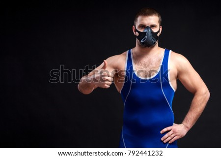 A young dark-haired man fighting Greco-roman wrestling and grappling in a blue   tights  , training mask holds the thumb up and posing against a black isolated  background