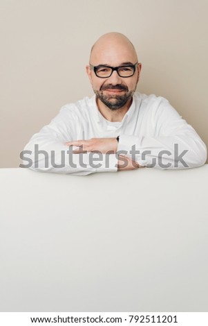 Smiling bald man in glasses leaning on white table with copy space