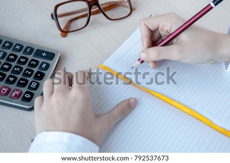 A girl in a white shirt writes in a notebook with a pencil, on the desk a calculator and glasses