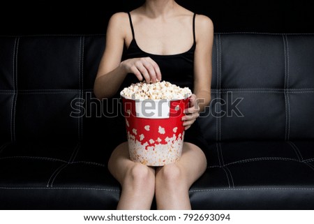 Girl sitting on a black leather couch holding popcorn box in hands