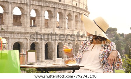 Happy young woman tourist drinking coffee and juice with cornetto at the table outside a bar restaurant in front of the Colosseum in Rome.