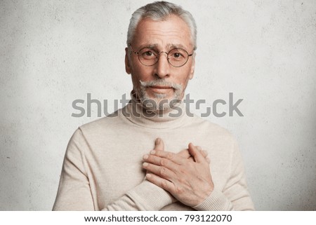 Portrait of kind friendly male pensioner feels lonely, expresses his love and good attitude to grandchildren, keeps hands on heart, proves that he tells only truth, isolated over white background Royalty-Free Stock Photo #793122070
