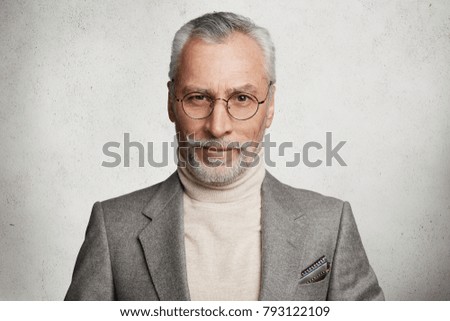 Portrait of positive pleased grey haired imposing male in formal wear and spectacles, expresses his confidence and self assuarance, poses against white concrete wall. Mature pensioner indoor Royalty-Free Stock Photo #793122109