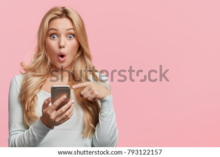 Horizontal shot of pretty young female with long hair looks with bugged eyes, holds modern smart phone, recieves unexpcted message from friend, reads reminder, isolated over pink background. Royalty-Free Stock Photo #793122157