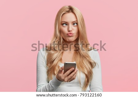 Crazy funny comic female crosses eyes, pouts lips, makes grimace, foolishes, holds cell phone, involved in games online, isolated over pink background. Facial expressions. Childish woman plays fool
