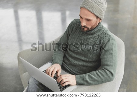 Top view of successful stylish male copywriter keyboards something on digital netbook, searches information on different websites, demonstrates productive work, sits in comfortable armchair.