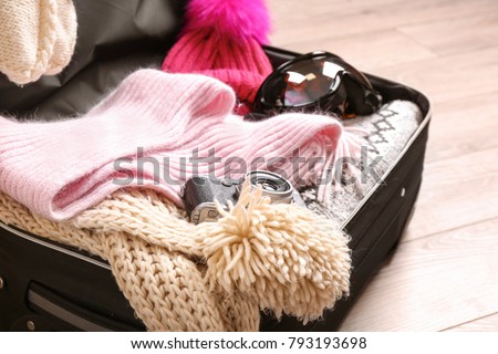 Open suitcase with warm clothes, photo camera and ski goggles on wooden floor. Winter vacation concept