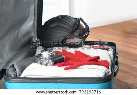 Open suitcase with warm clothes, photo camera and ski outfit on floor. Winter vacation concept